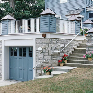 A blue garage door set into a stone wall, with a railed deck above and concrete stairs leading up to the house entrance.