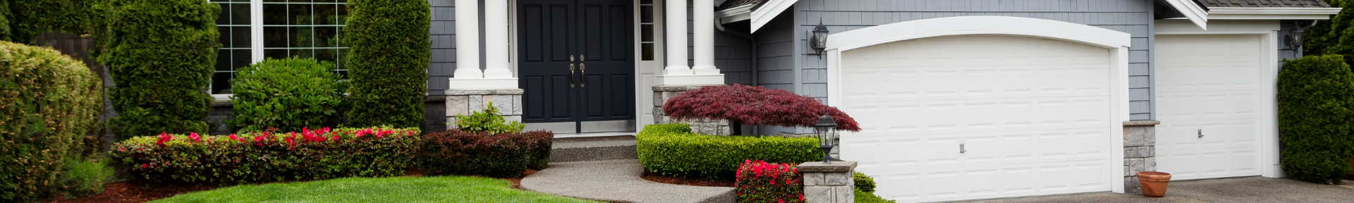 A grey house with a blue front door, white pillars, a wide white garage door, and a neatly manicured lawn with shrubs.