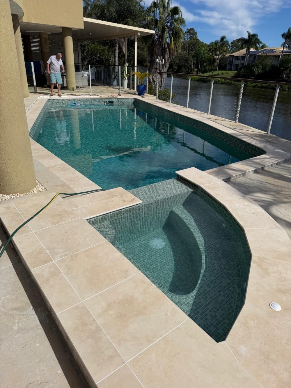 Backyard swimming pool with a curved shallow area, beige tile deck, and waterfront view