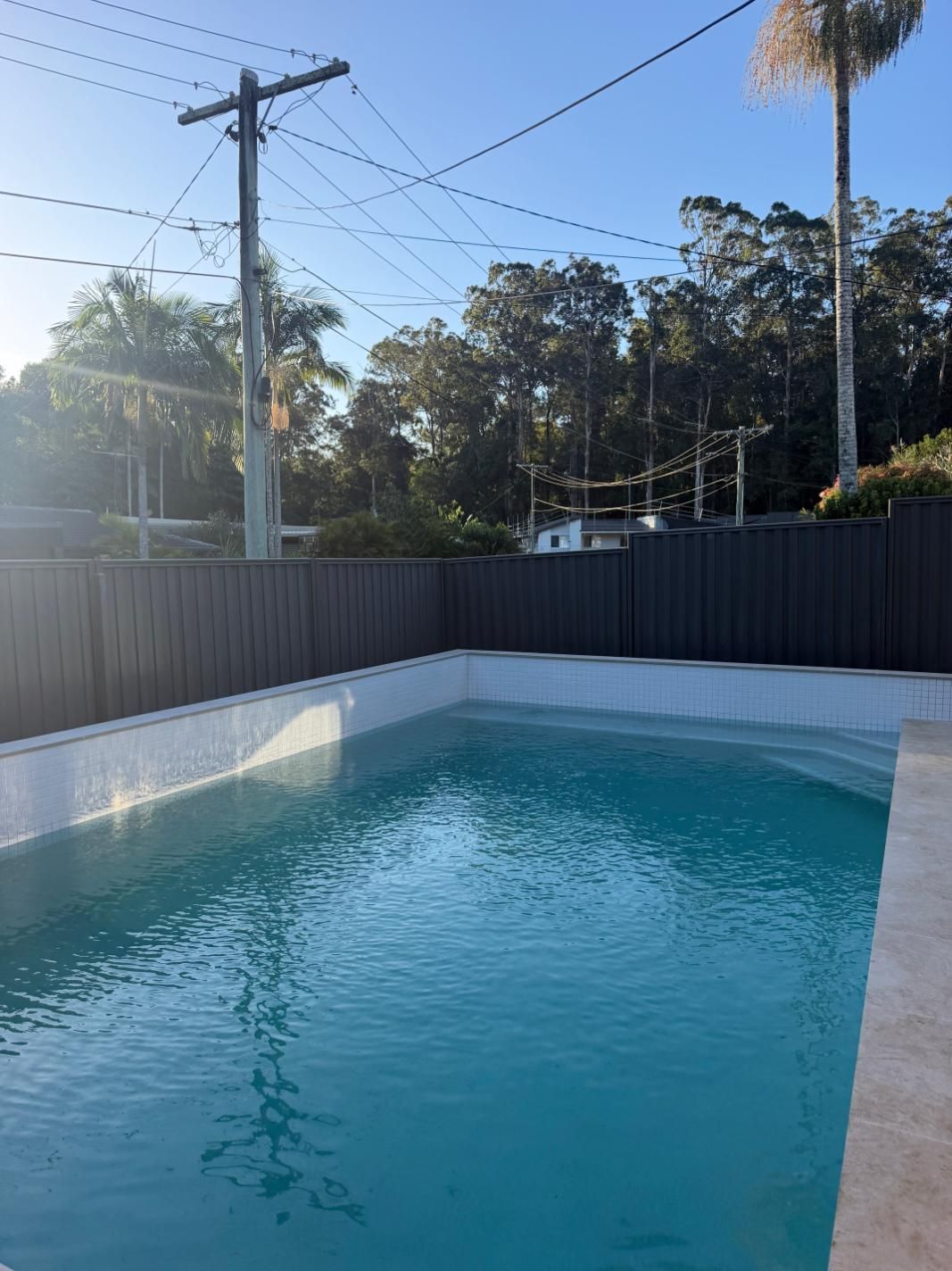 Backyard swimming pool with clear blue water, white walls, and tall fence under a sunny sky