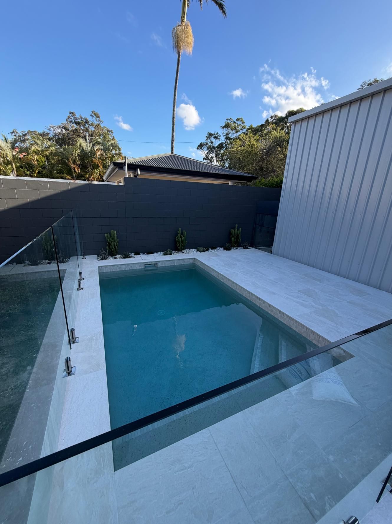 Modern backyard pool with clear water, white deck, dark fence, and blue sky.