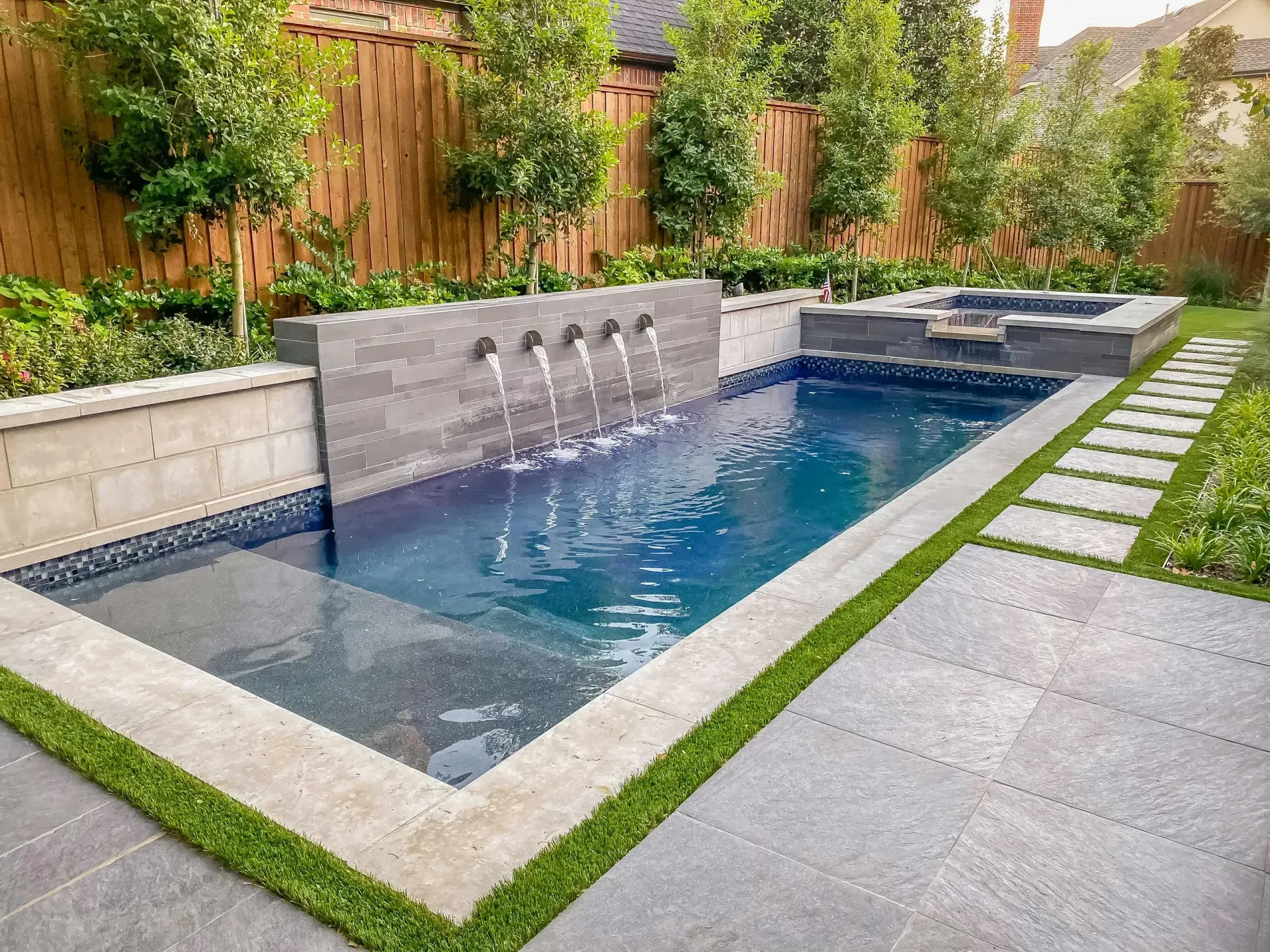 A rectangular pool with a fountain and spa. Features gray stone and green grass, with a wooden fence background.