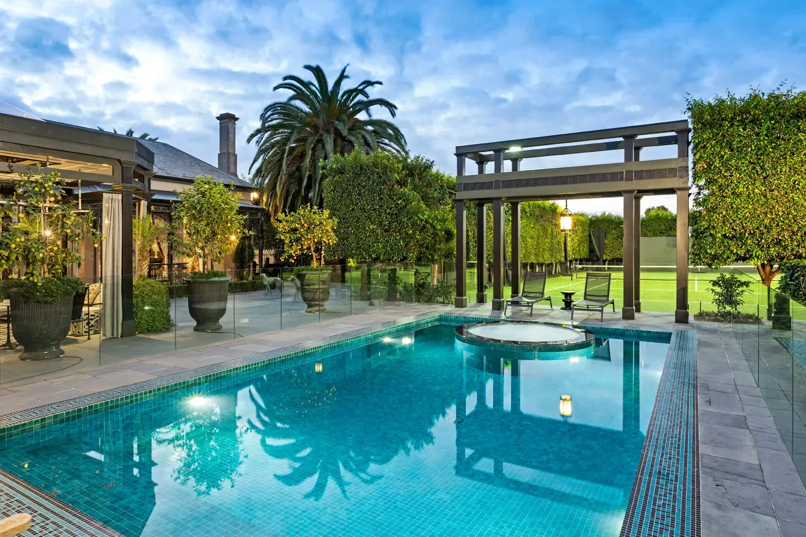Swimming pool surrounded by greenery, featuring a pergola, with a house in the background.