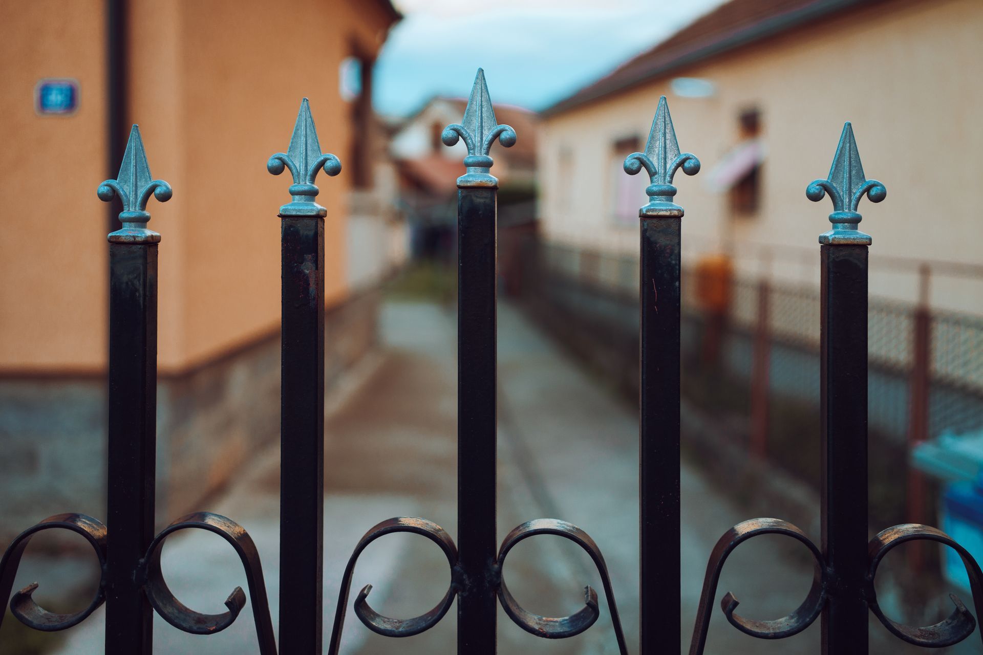 Black metal fence with decorative fleur-de-lis tops, blurred background of houses and a road.