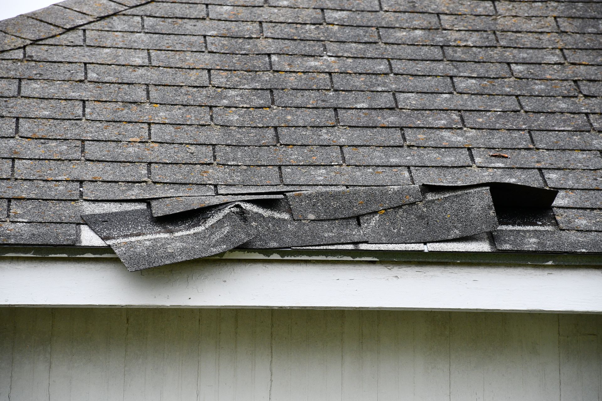 Damaged asphalt roof shingles, torn and curled, over a white-painted building.