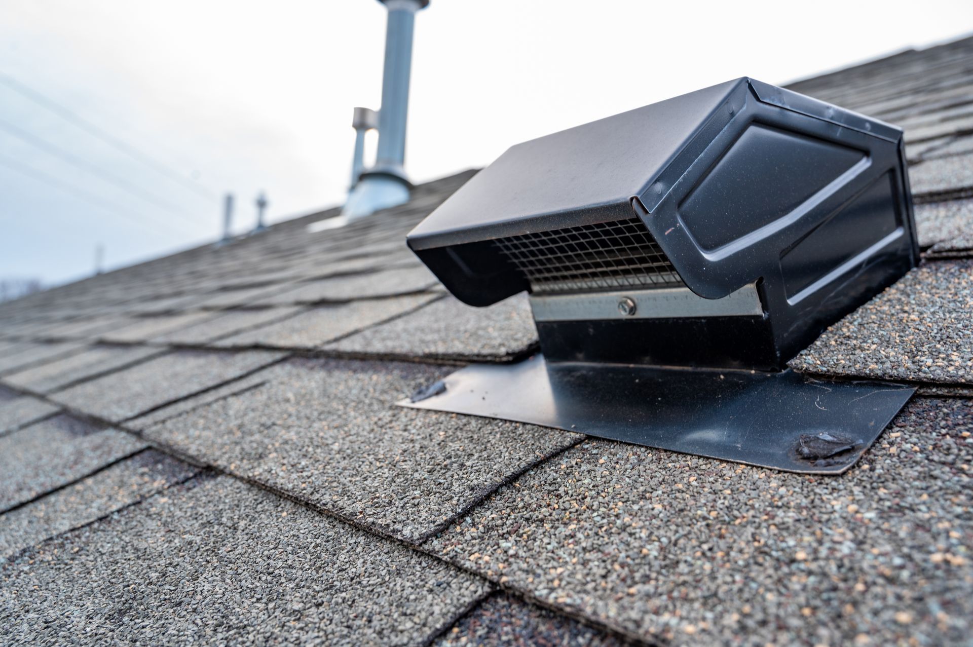 Black roof vent on a gray shingle roof. Steel chimney in the background. Cloudy sky.
