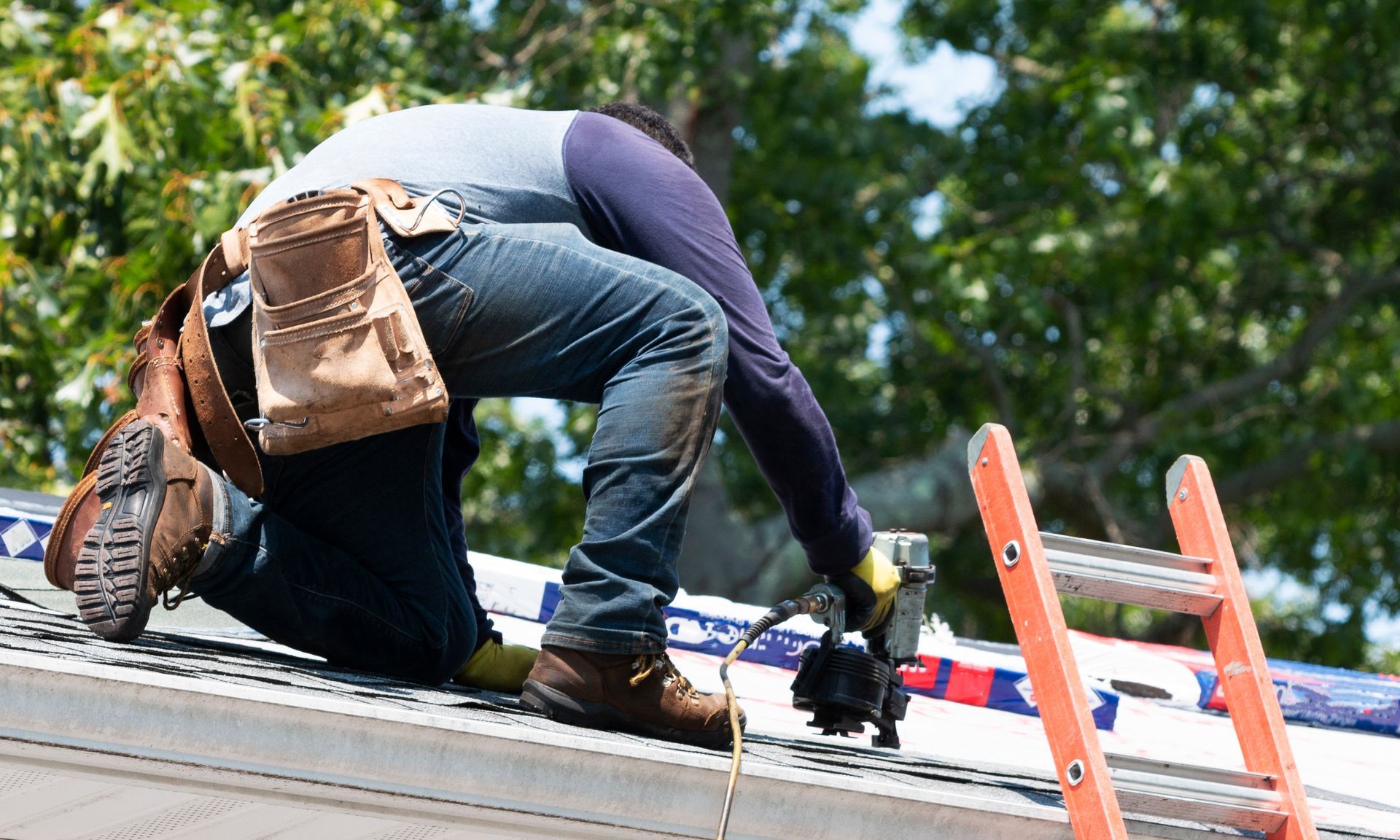 Roofer kneeling on a roof, using a nail gun. A ladder is propped nearby.