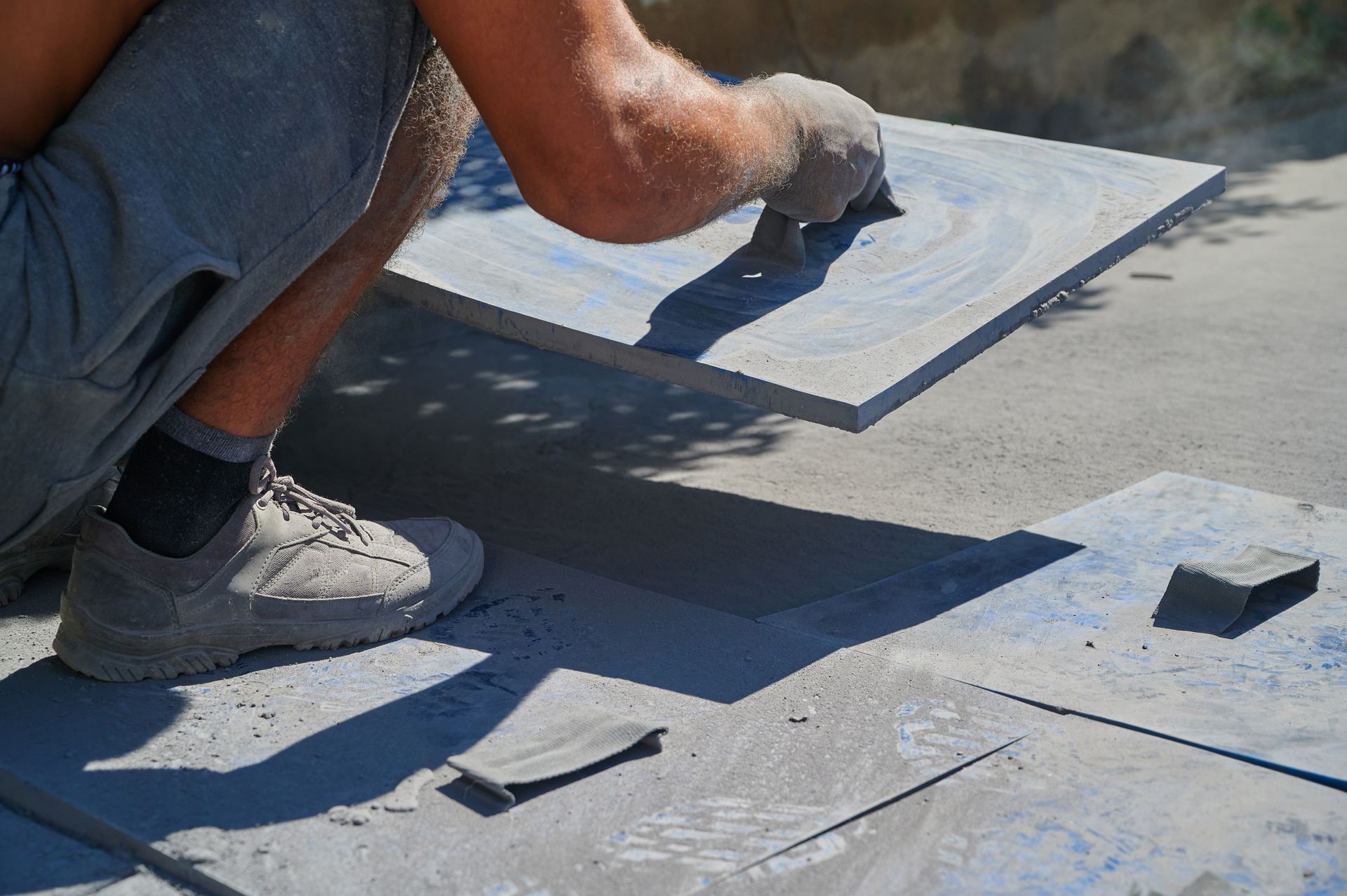 Person installing a tile, using a trowel. Outdoors, tiles are gray and concrete is visible.