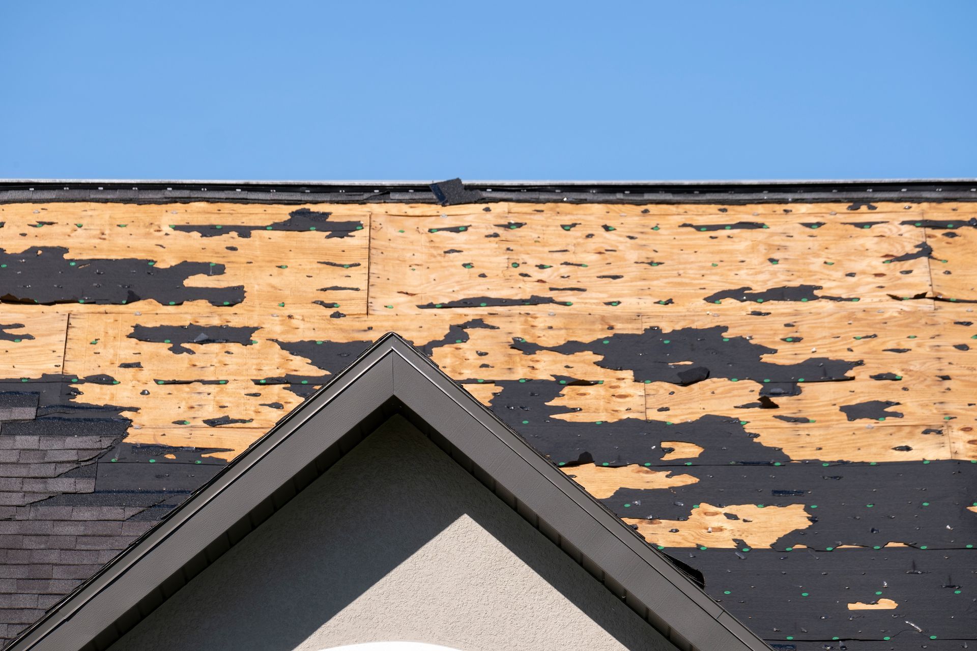 Roof with missing shingles revealing wood panels and black underlayment against a blue sky.