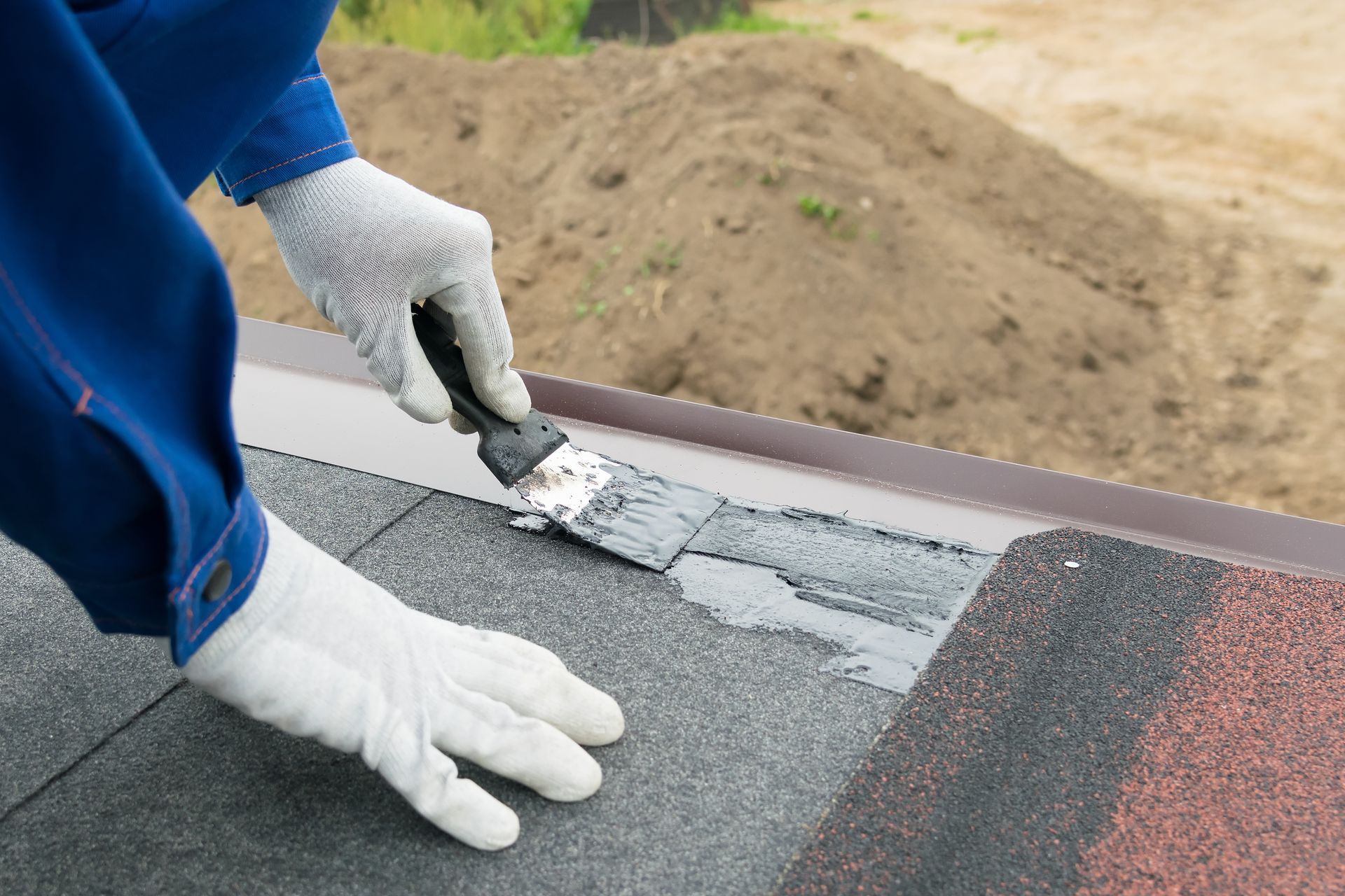 Person in blue overalls and gloves applying sealant to a dark asphalt shingle roof with a trowel.