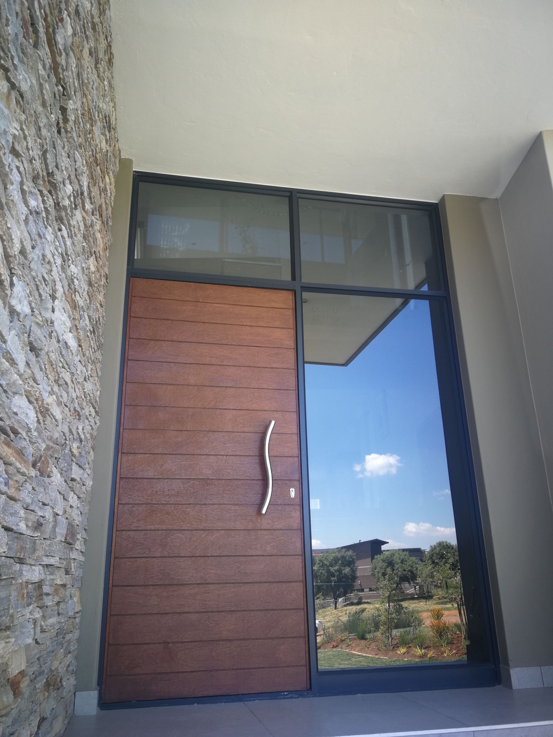Wooden front door with glass panel, stone wall to the left, sky visible through the glass.