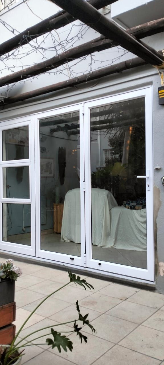 White framed glass doors, partially open, lead to a room with a draped table. A pergola and plants are in the foreground.