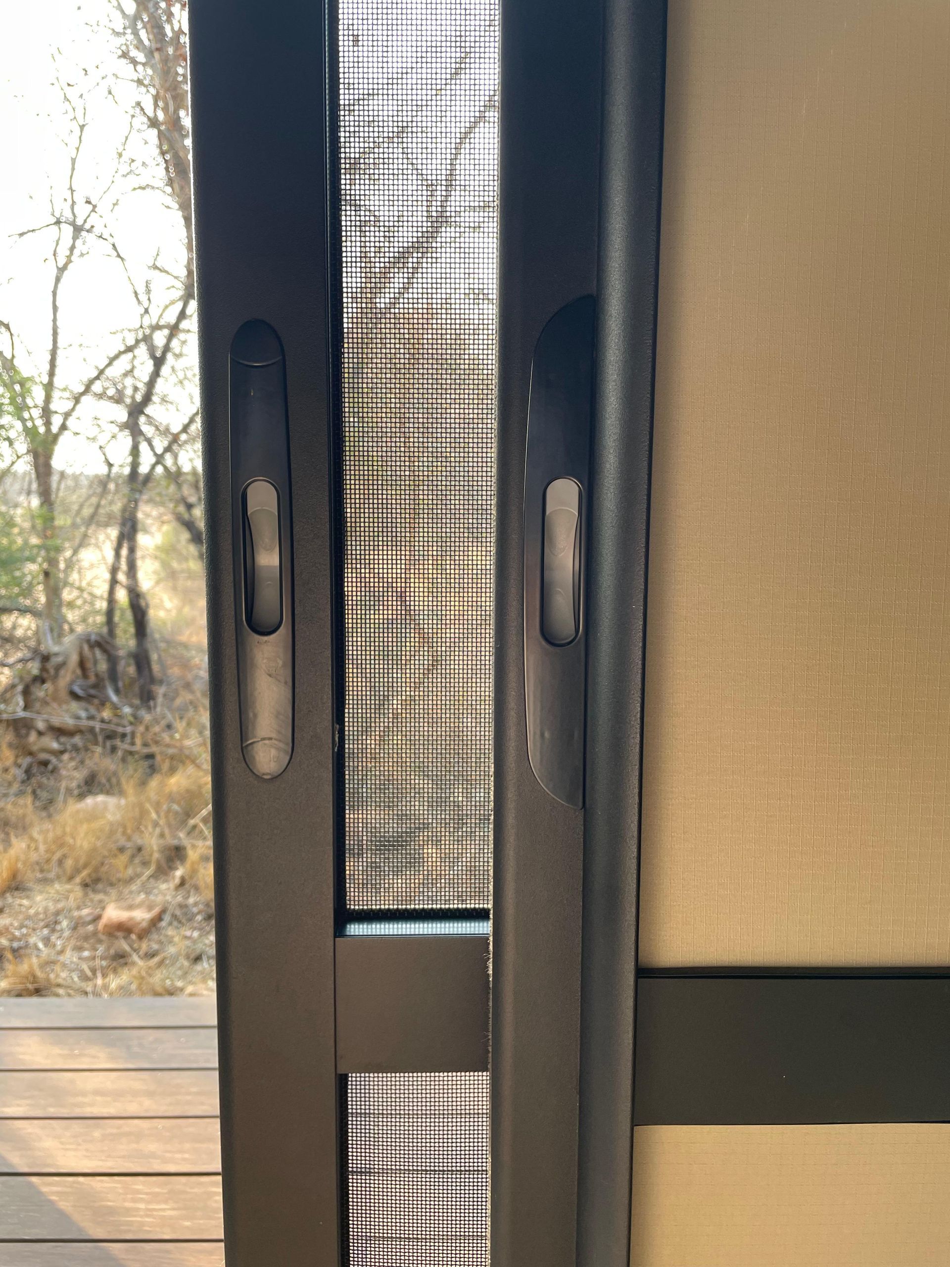 Screen door, closed. Dark frame with mesh screen. Handles on each side. Outdoor view.