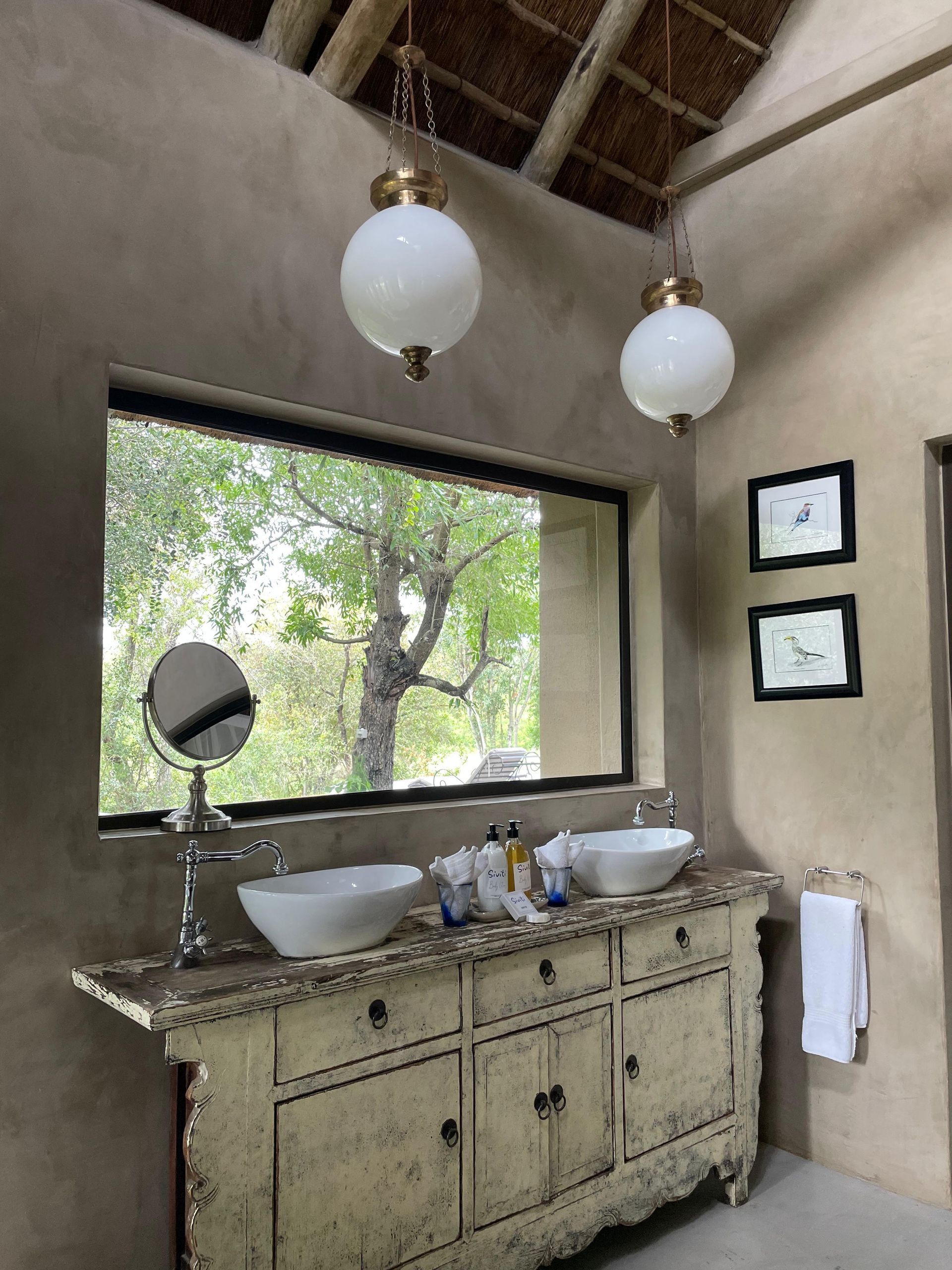 Bathroom with distressed white vanity, two white bowl sinks, window overlooking trees, two globe pendant lights.