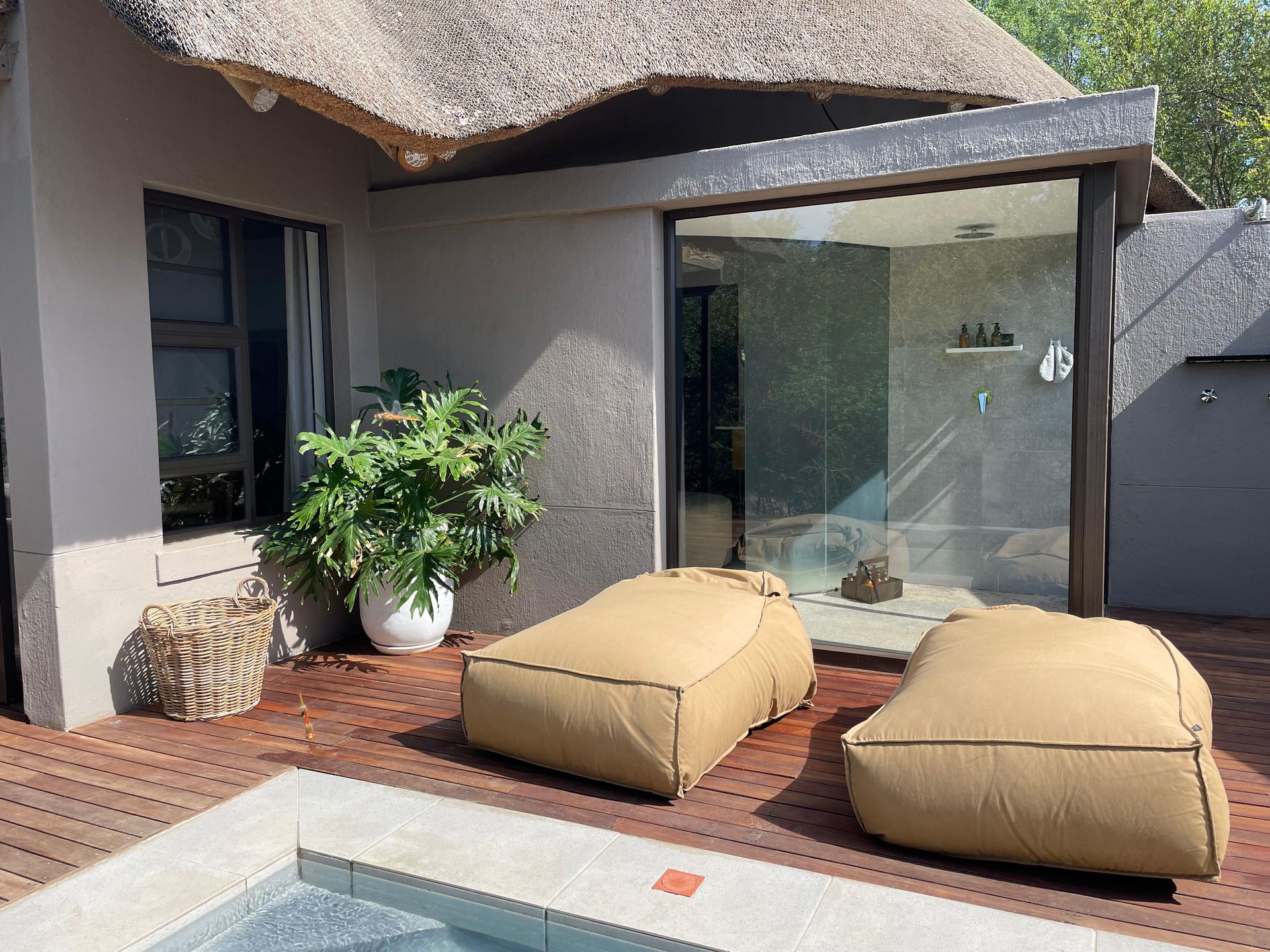 Patio with two tan beanbag chairs, a pool, and a building with a thatched roof and glass shower.