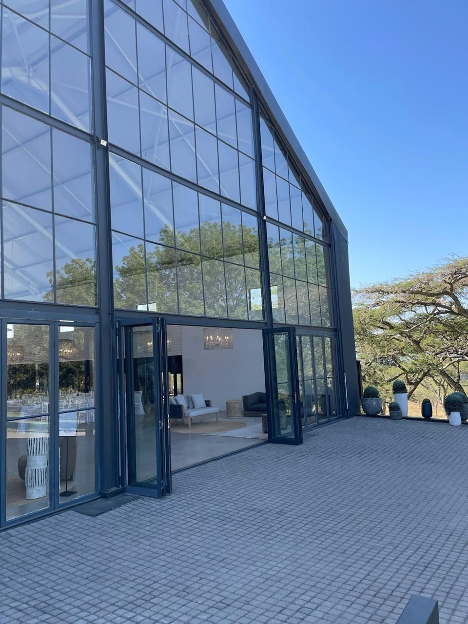 Glass-walled event space with open doors, overlooking a paved patio. Sunny day, trees visible.