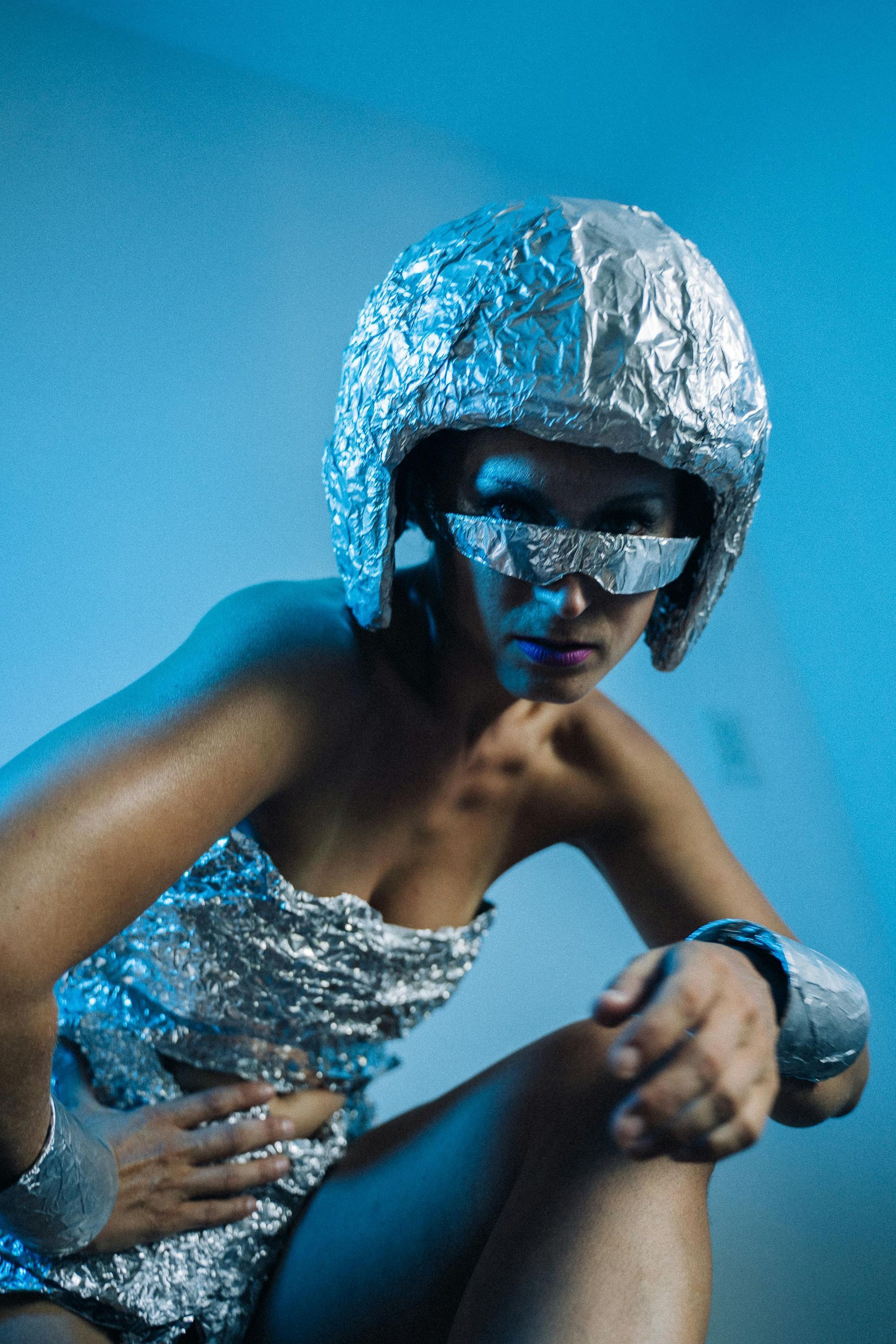 Woman wearing a foil helmet, eye shield, strapless top, and bracelets, poses with a blue background.