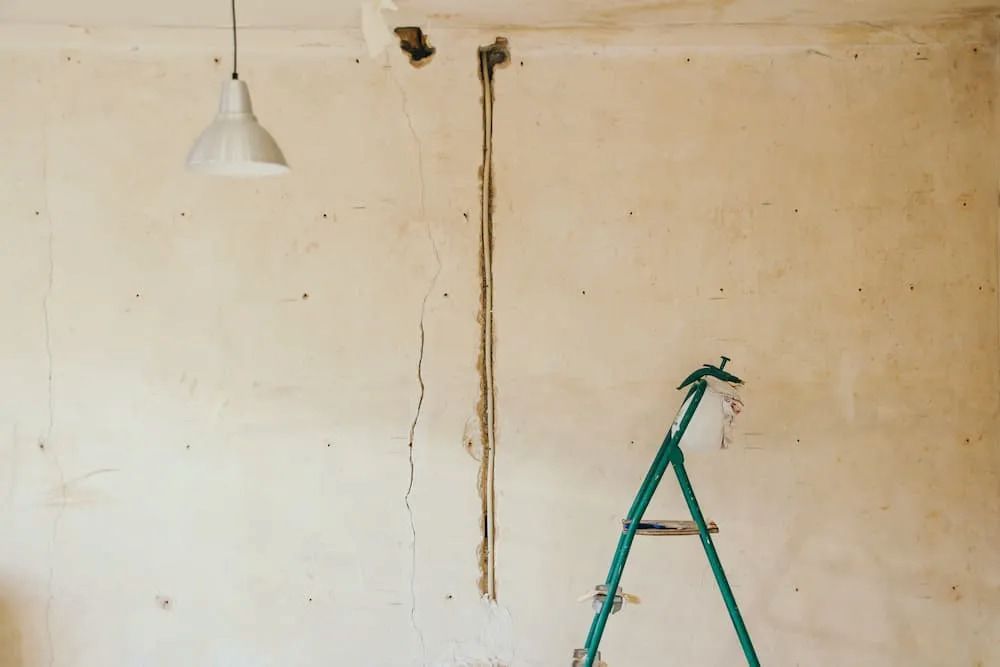Cream-colored wall with long crack, exposed wiring, a white lamp, and a green ladder. Construction site. — Unique Waterproofing Solutions in North Rocks, NSW