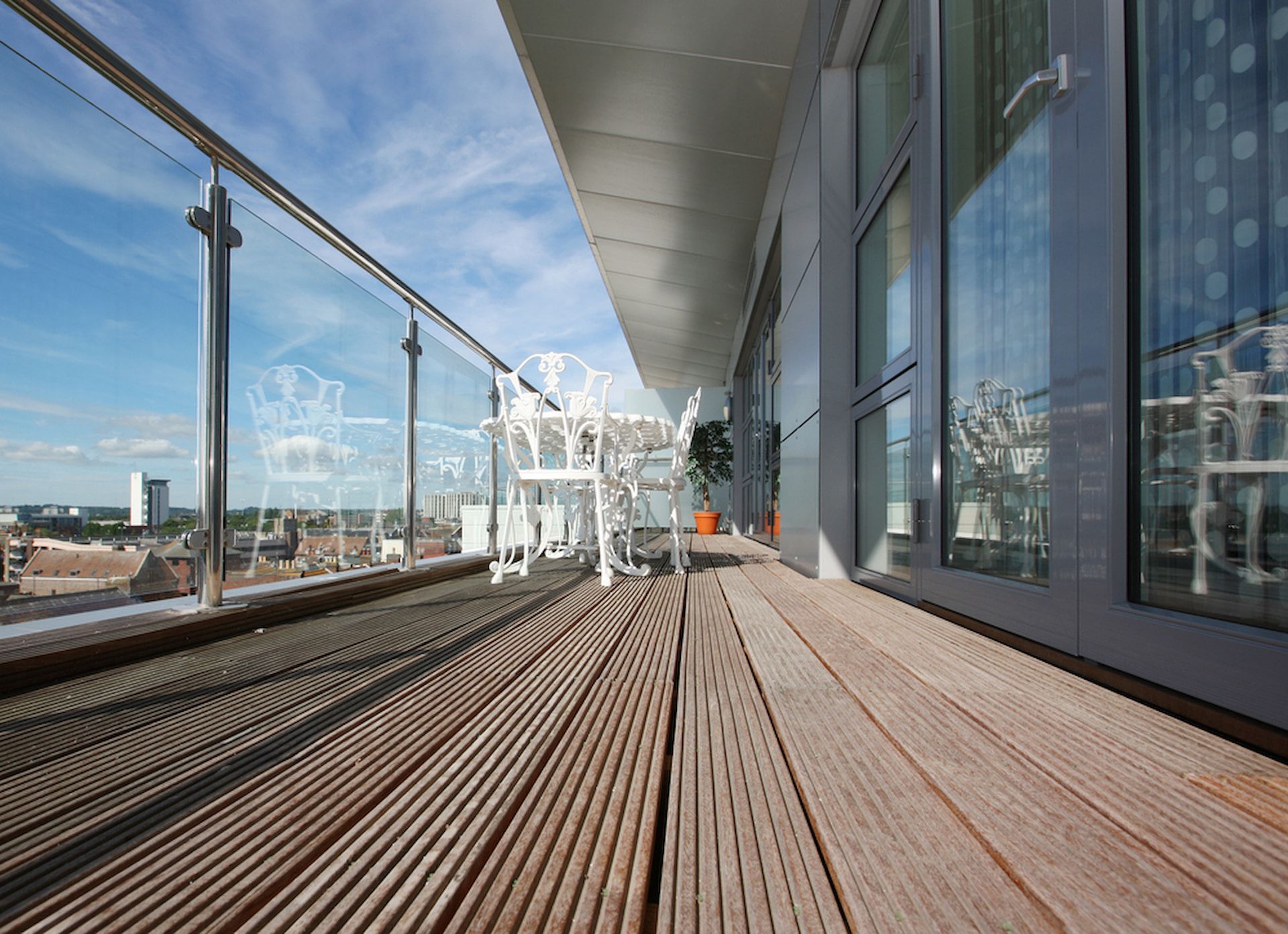 Wooden balcony with glass railing, chairs, and city view under a blue sky. — Unique Waterproofing Solutions in North Rocks, NSW