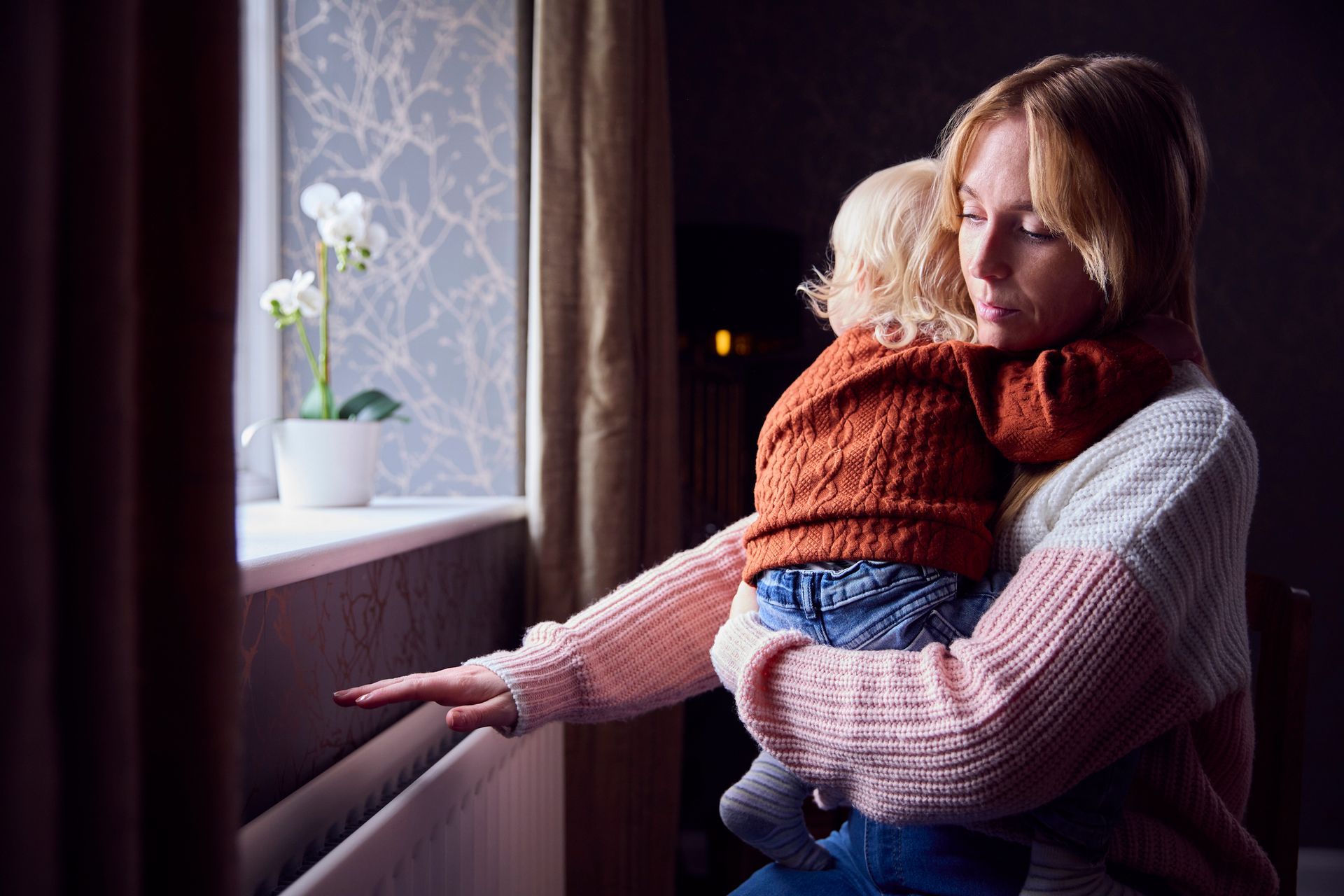 Mother With Son Trying To Keep Warm By Radiator At Home Because Of Broken Heater.