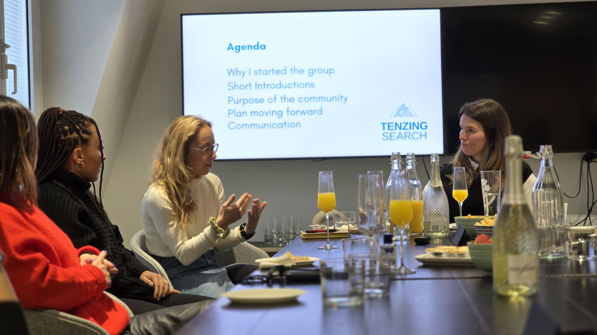 People sit around a table in a meeting room, facing a screen showing an agenda under the heading "Leading Search."