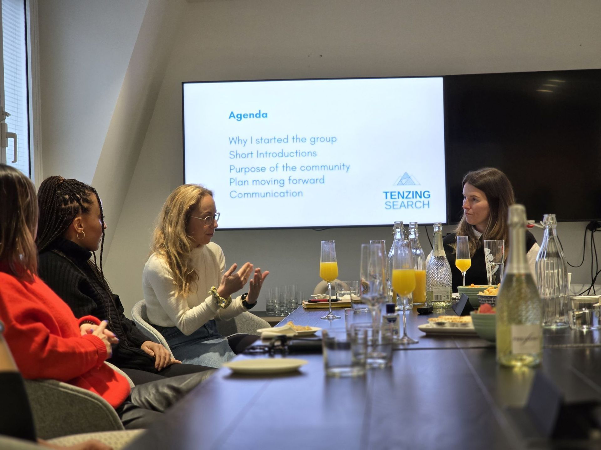 A presentation in a modern office with an audience seated, facing three speakers seated in front of a digital screen.