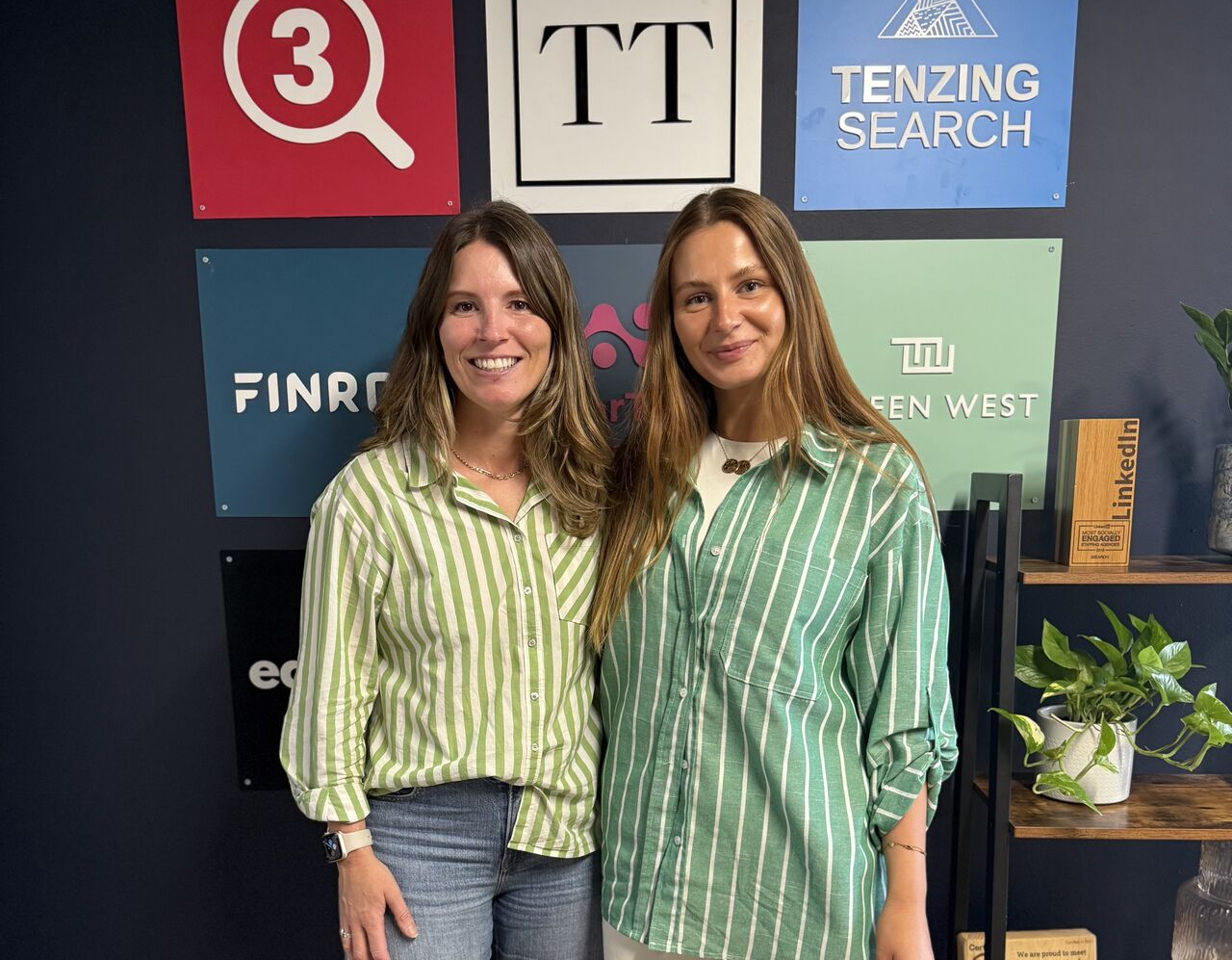 Two people stand side-by-side smiling in front of a wall featuring various company logos and a plant on a shelf.