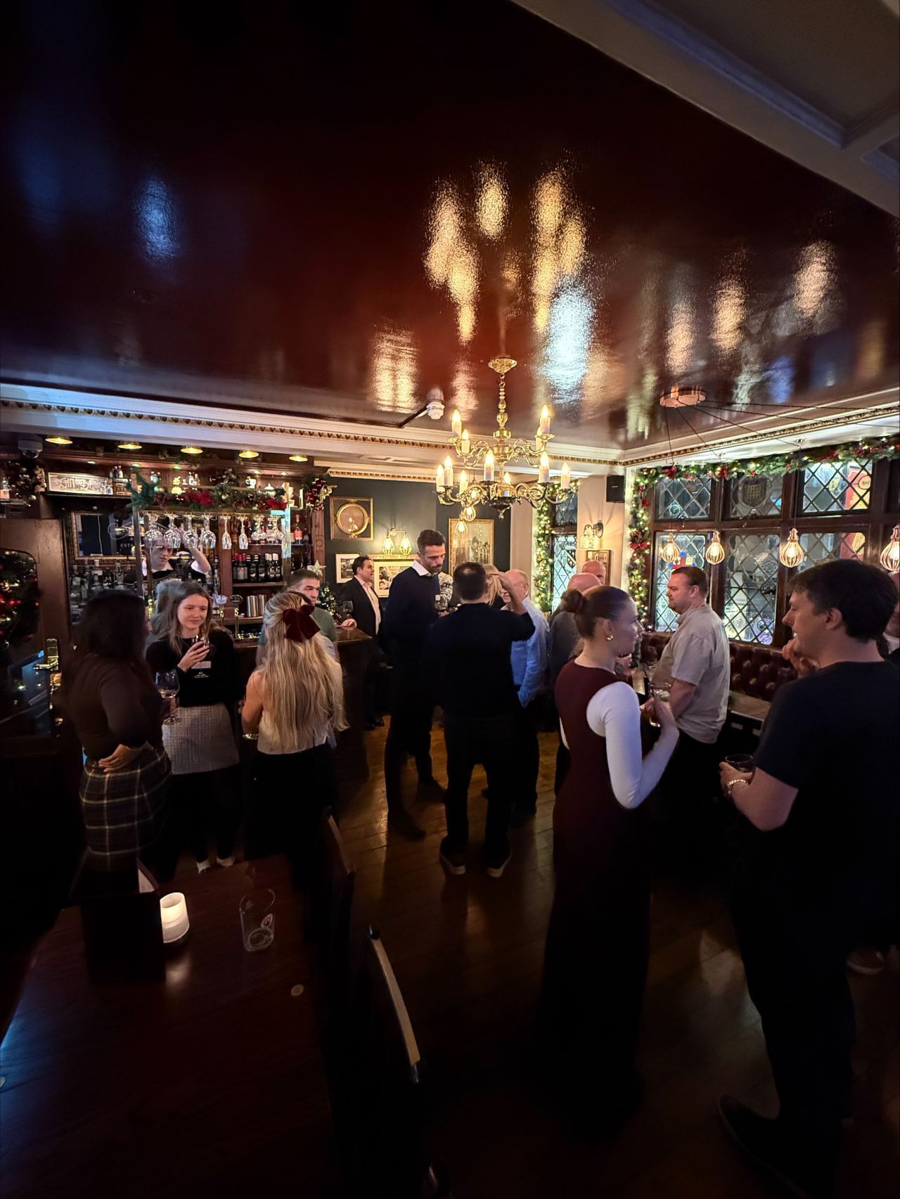 Three people talk in a brightly lit office setting, with a woman in a black blazer holding a glass while conversing.