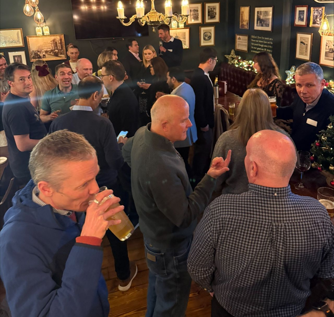 Three people talk in a brightly lit office setting, with a woman in a black blazer holding a glass while conversing.
