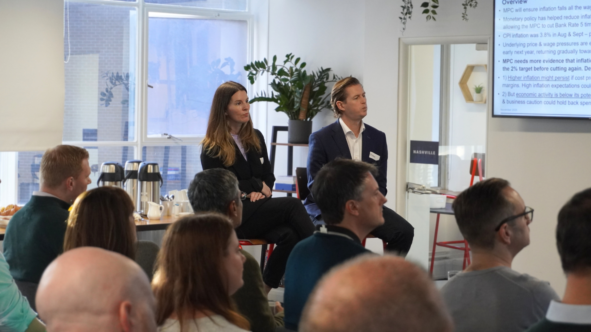 Three people talk in a brightly lit office setting, with a woman in a black blazer holding a glass while conversing.