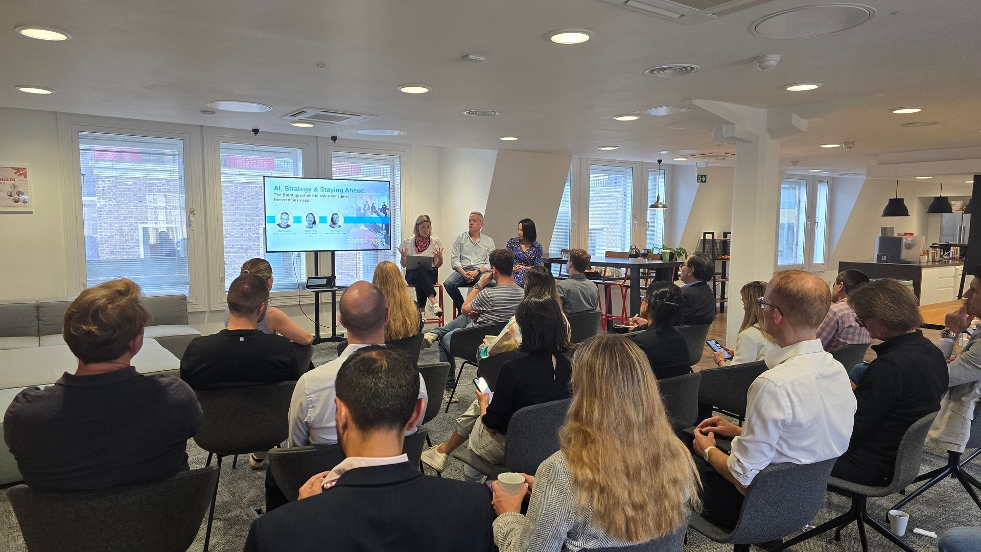 Three people talk in a brightly lit office setting, with a woman in a black blazer holding a glass while conversing.