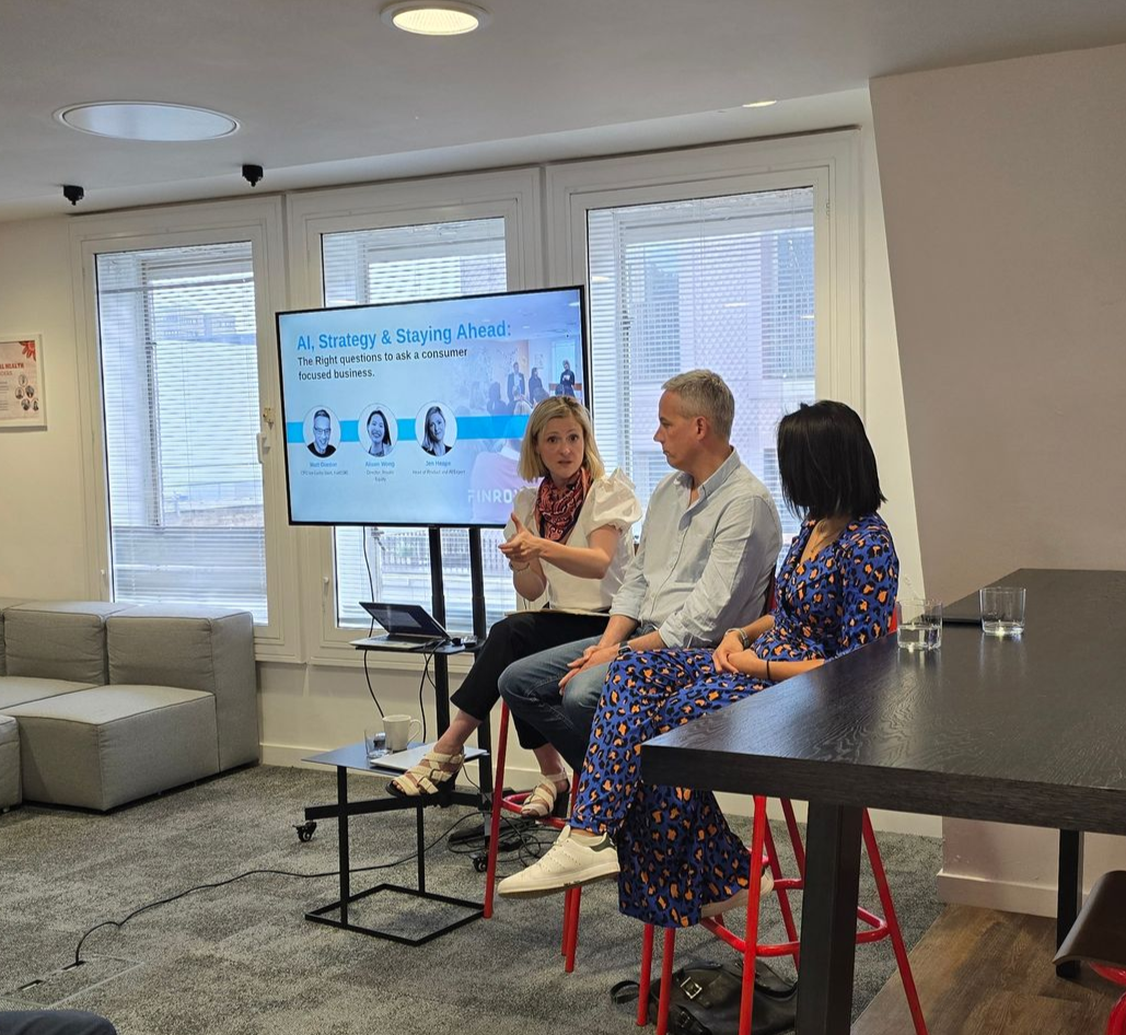 Three people talk in a brightly lit office setting, with a woman in a black blazer holding a glass while conversing.