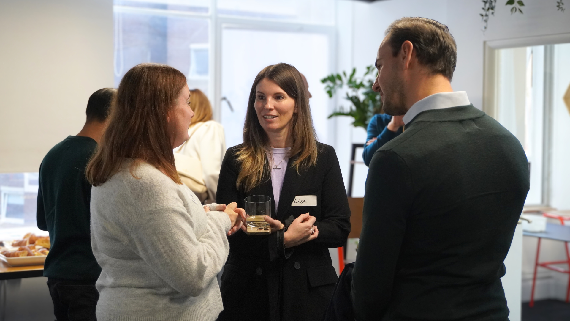 Three people in a bright office space converse while standing near a desk with refreshments.