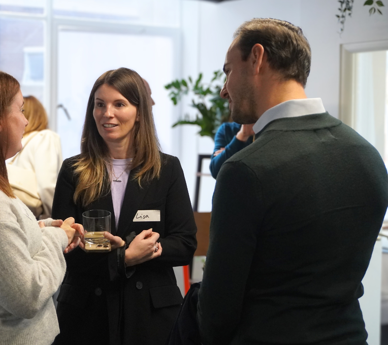 A person in a black blazer with a name tag speaks to others while holding a drink during a professional networking event.