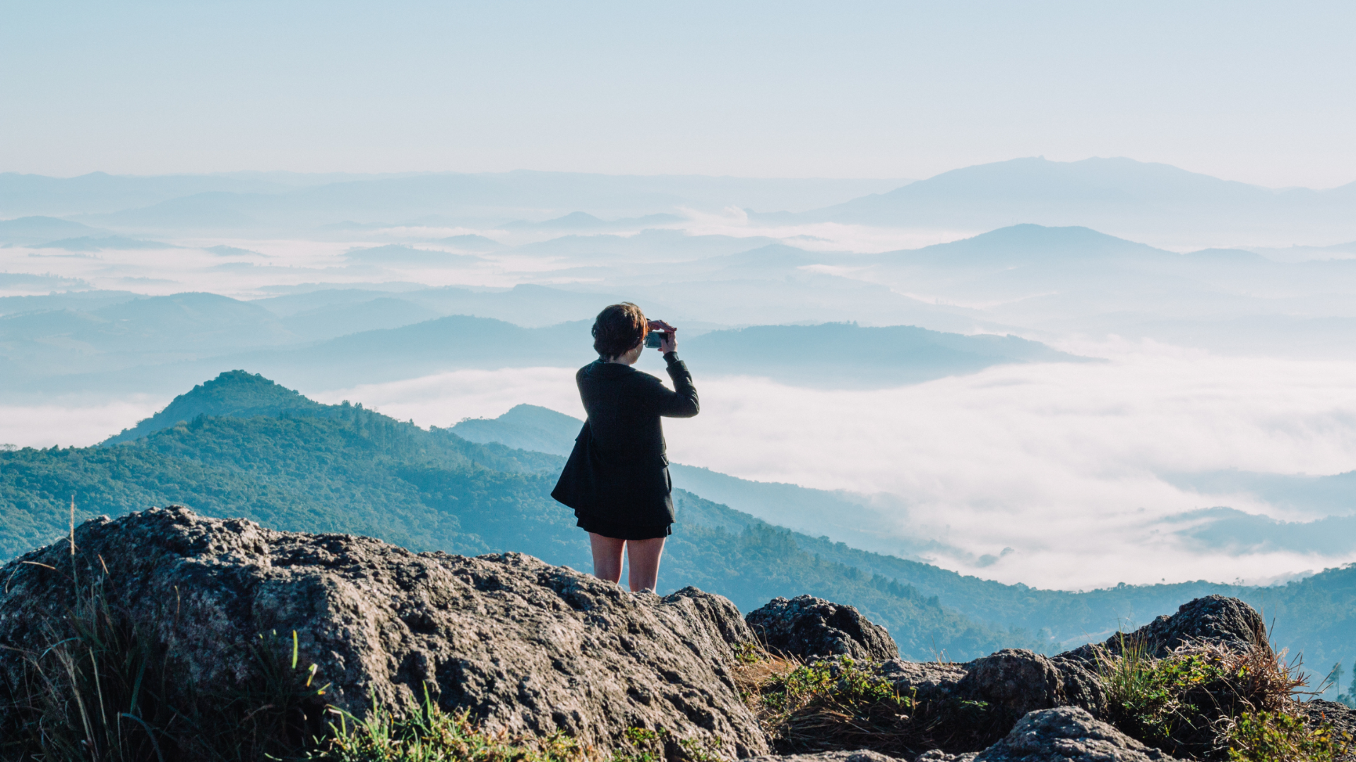 A person stands on a rocky mountain summit, looking through binoculars at a vast, misty mountain landscape at dawn.