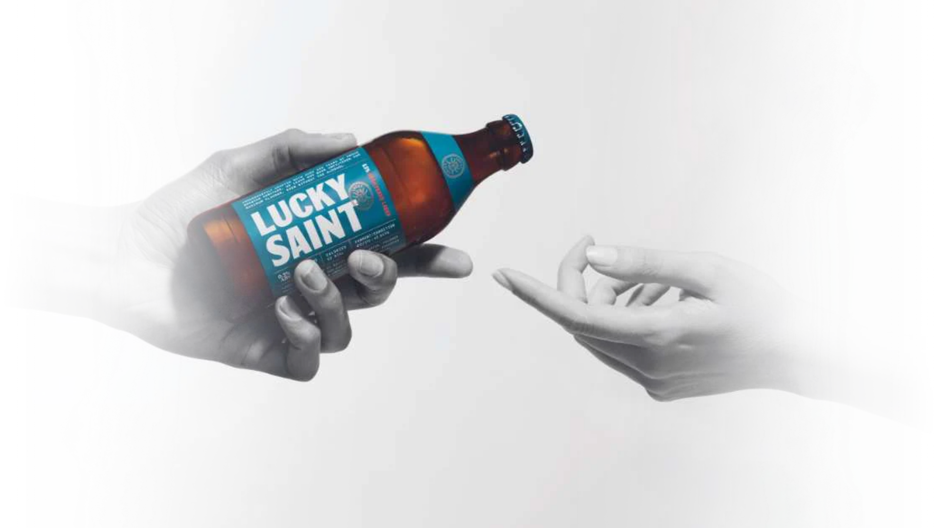 A black-and-white image shows one hand offering a bottle of Lucky Saint beer to another outstretched hand.