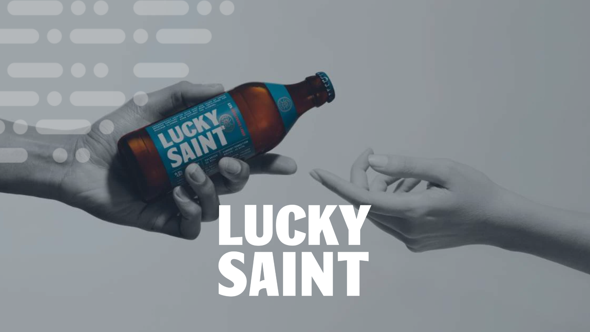 Two hands meet against a grey background, one holding a Lucky Saint non-alcoholic beer bottle.