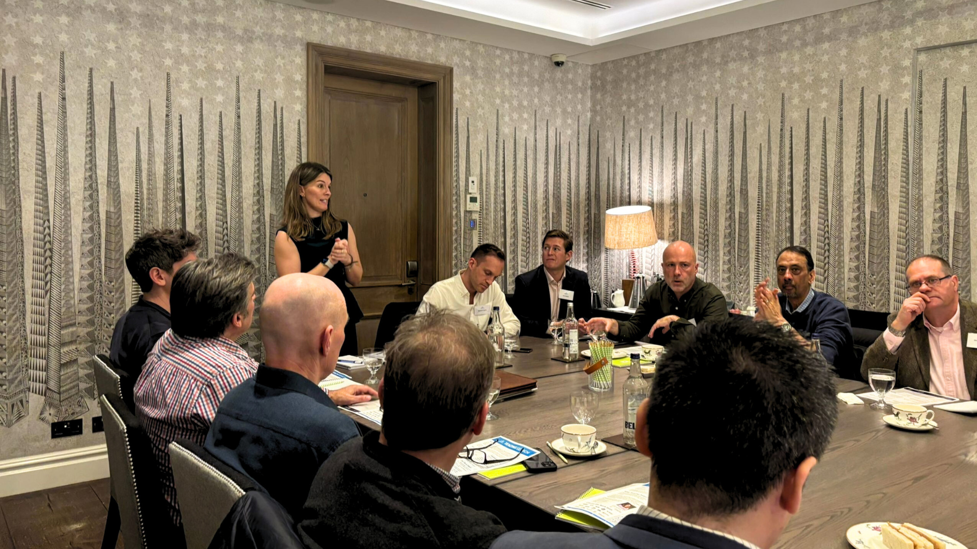 Three people talk in a brightly lit office setting, with a woman in a black blazer holding a glass while conversing.