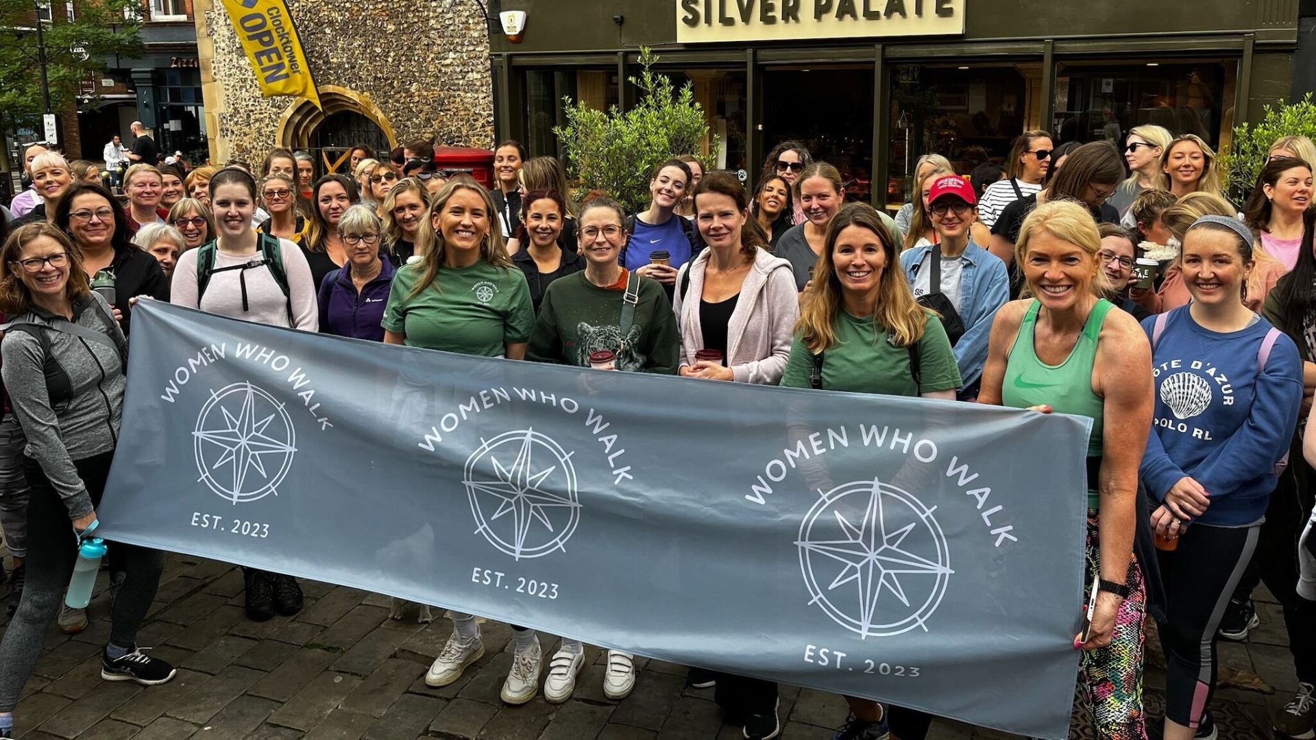 Two people stand side-by-side smiling in front of a wall featuring various company logos and a plant on a shelf.