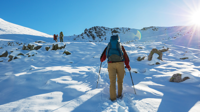 A hiker with a blue backpack and trekking poles walks through deep, sunlit snow toward others on a mountain slope.