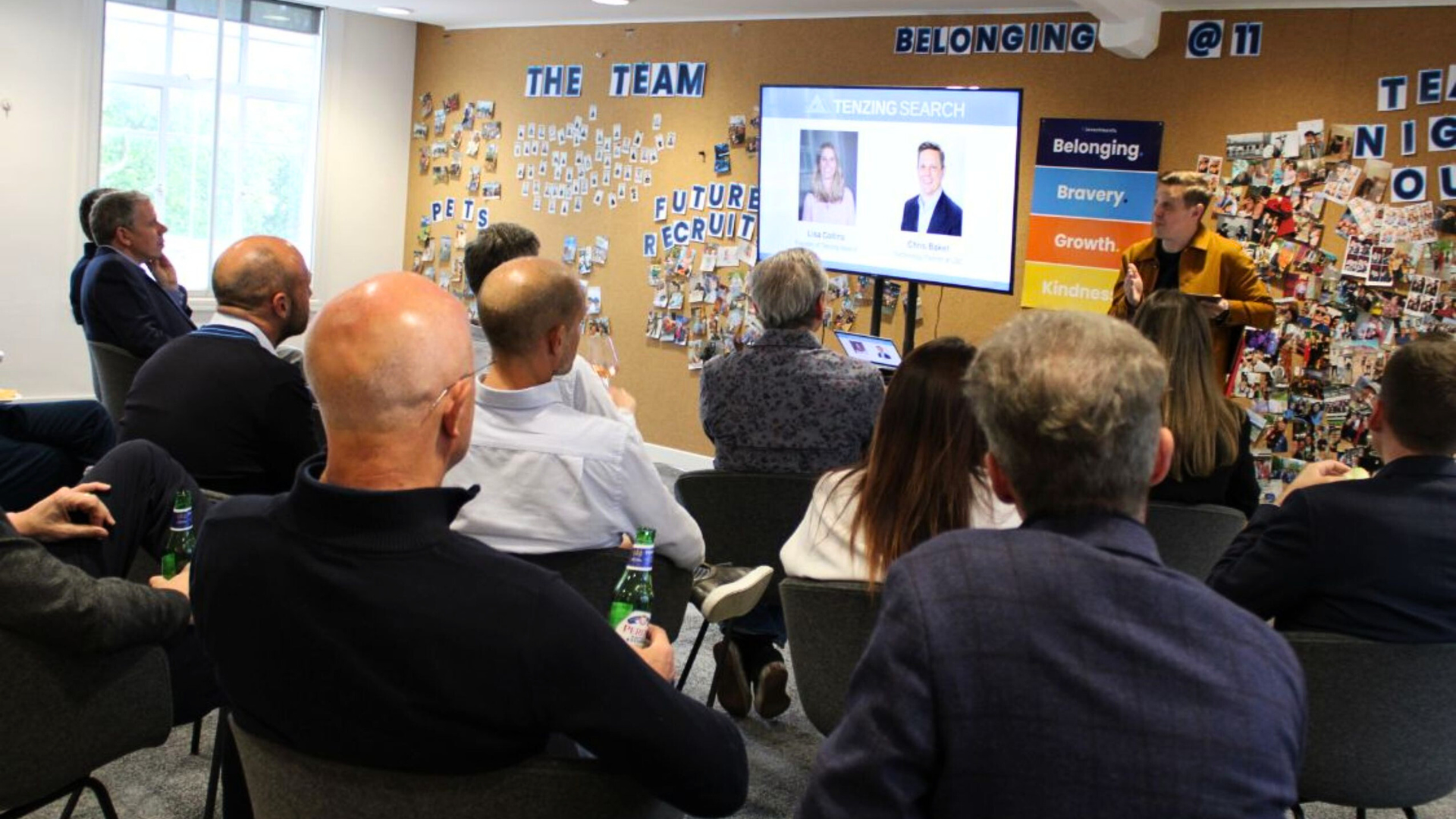 Three people talk in a brightly lit office setting, with a woman in a black blazer holding a glass while conversing.