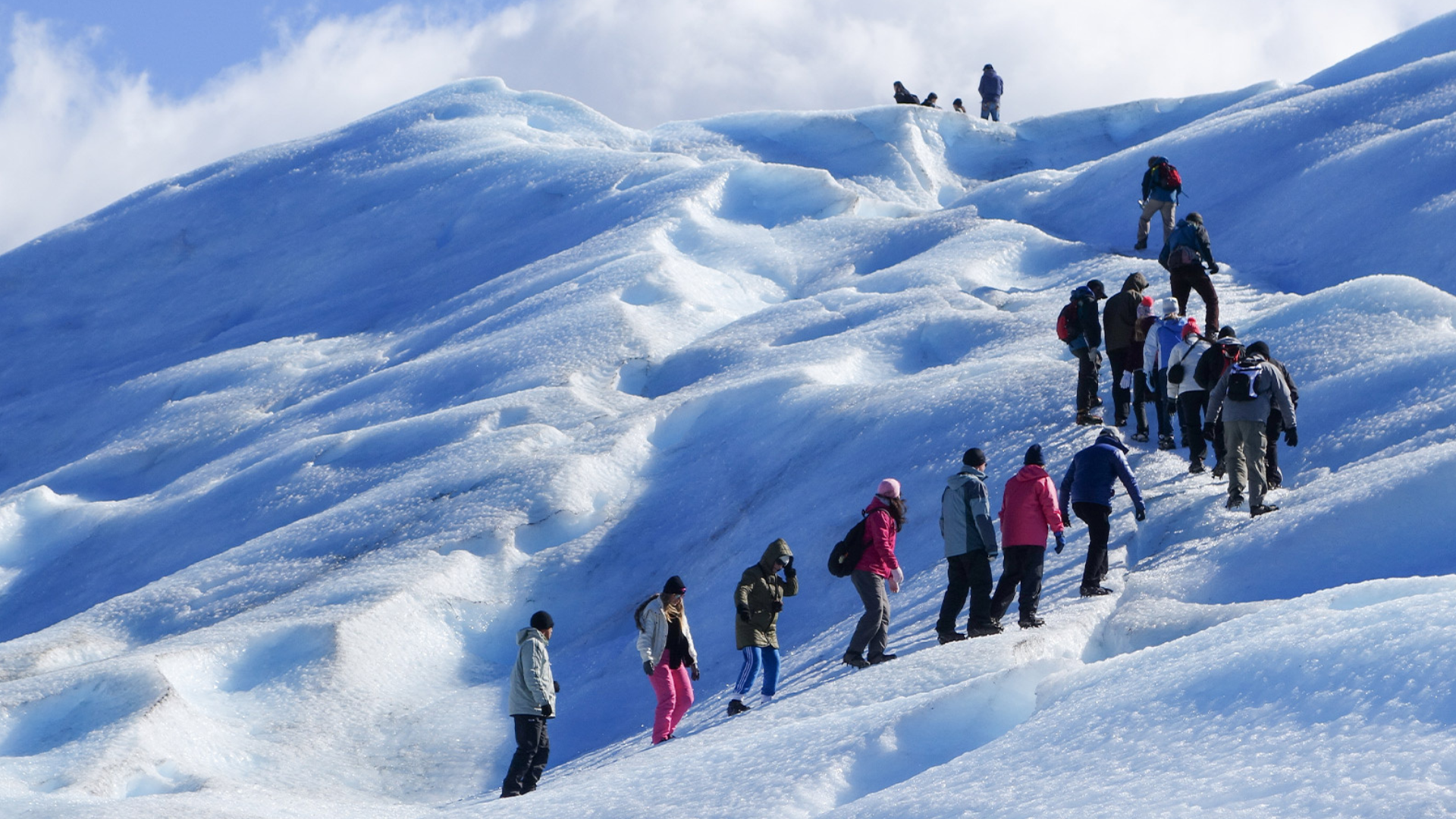 A group of people wearing winter clothing hiking up a steep, sunlit blue glacier.