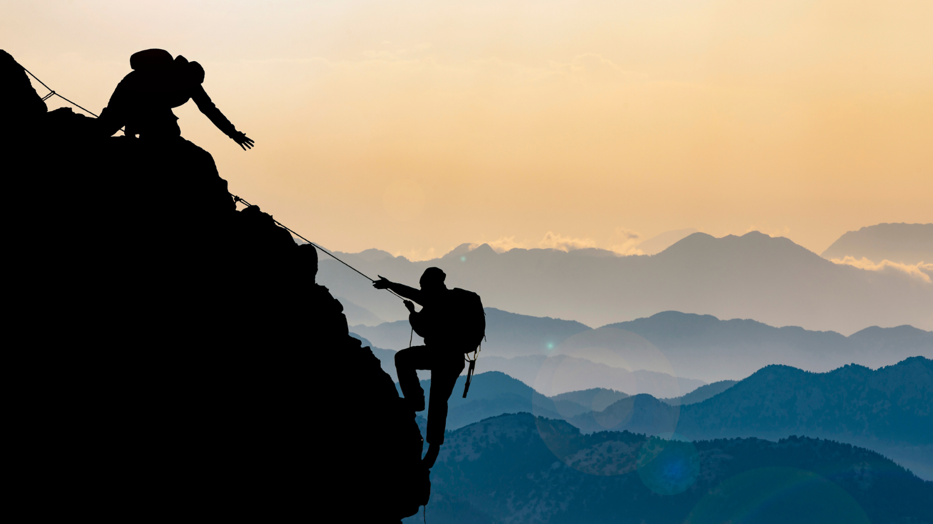 Two hikers in silhouette climb a steep, rugged mountain ridge against a hazy, golden mountain range at sunset.