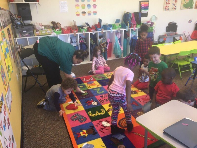 Children and an adult in a classroom play on a colorful rug with numbers.