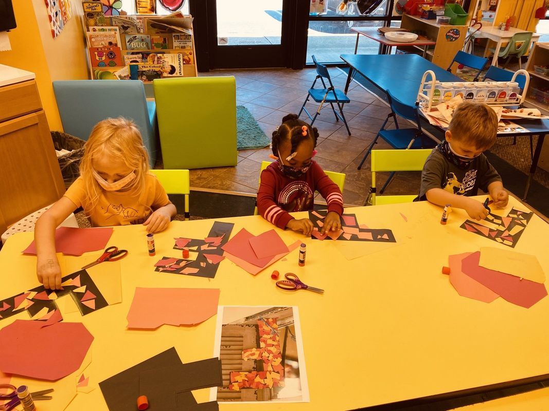 Children craft at a yellow table. They are wearing masks. There are construction papers and supplies on the table.