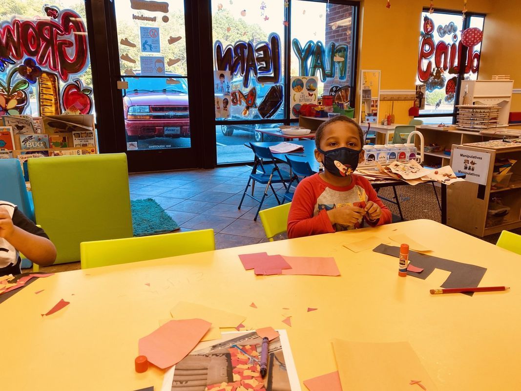 Child at a table in a classroom wearing a mask, doing arts and crafts.