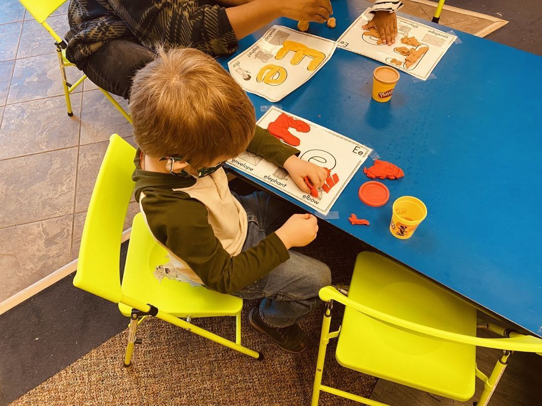 A child sits at a blue table, using clay. Yellow chairs. Another person works at the table.