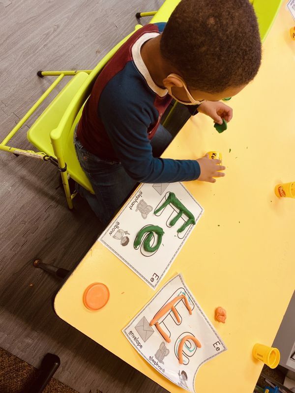 Boy with mask molds clay letters on a yellow table with letter guides.