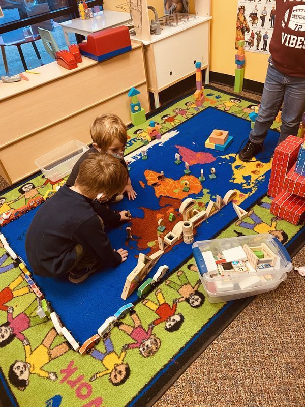 Two children playing with blocks on a world map rug in a classroom.