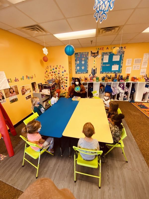 Children and a teacher sit around a blue/yellow table in a brightly colored classroom.